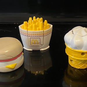 May include: Three toy fast food items: a hamburger, a container of fries, and an ice cream cone. The hamburger is beige with red and yellow accents. The fries are yellow and in a white container with the McDonald's logo. The ice cream cone is yellow and white.