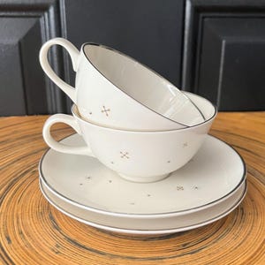 May include: Two white teacups with gold star and cross designs and a silver rim. The cups are stacked on top of each other and are sitting on a white saucer with a silver rim.