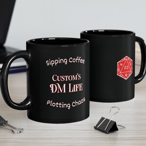Könnte beinhalten: Zwei schwarze Kaffeetassen mit weißem Design. Die Tasse links trägt die Aufschrift "Sipping Coffee Custom's DM Life Plotting Chaos". Die Tasse rechts hat ein rotes 20-seitiges Würfel-Motiv.