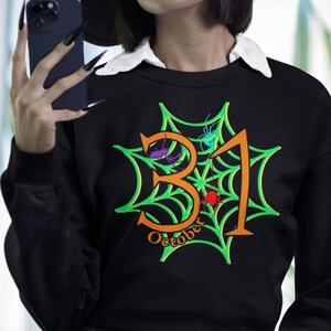 May include: Black sweatshirt with a green spiderweb design and the text "October 31" in orange. The sweatshirt has a white collar and is worn by a person wearing a black witch hat.