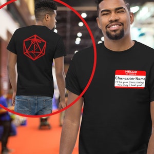 Puede incluir: Camiseta negra con un símbolo médico rojo dentro de un dado rojo de veinte caras en la espalda. La parte delantera de la camiseta tiene una etiqueta de nombre roja que dice "HELLO MY NAME IS Character Name I'll be your Cleric today! How may I heal you?"