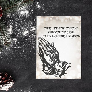 May include: A white card with a black and white illustration of praying hands. The text on the card reads "May divine magic surround you this holiday season."