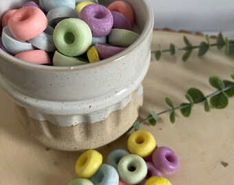 May include: A white ceramic bowl filled with pastel-colored donut-shaped candies. A small pile of the same candies is scattered on a light wood surface.
