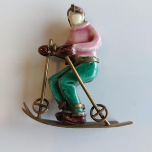 Può includere: Una figurina vintage in smalto di uno sciatore con un maglione rosa, pantaloni verdi e stivali marroni. Lo sciatore tiene bastoni da sci ed è su sci con ruote metalliche.