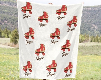 May include: A white blanket with a repeating pattern of red roller skates with tan detailing and the name "Joyce" in a cursive font. The blanket is displayed in a field with a mountain backdrop.