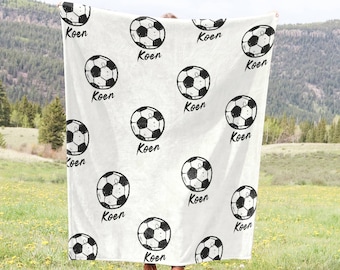 Puede incluir: Manta blanca con un patrón de balón de fútbol blanco y negro. La manta tiene el nombre "Koen" repetido en todo el diseño.
