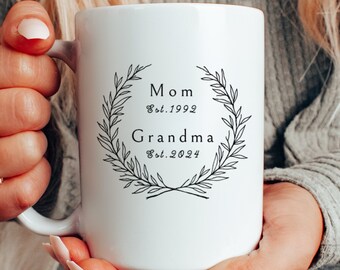 May include: White ceramic mug with a black design that reads "Mom Est. 1992 Grandma Est. 2024" surrounded by a wreath of leaves.