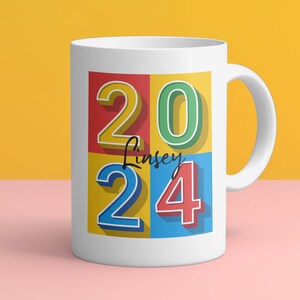 May include: White ceramic mug with a colorful design featuring the numbers 2024 in a retro style. The name "Lindsey" is written in black script on the mug.