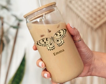 May include: A clear glass tumbler with a wooden lid and a straw, filled with a light brown beverage. The tumbler features a butterfly design and the name "Emma" in a script font. The tumbler is held by a person.
