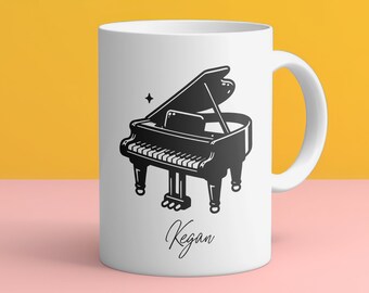 May include: White ceramic mug with a black and white illustration of a grand piano and the name "Kegan" written in cursive.