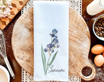 May include: White kitchen towel with a watercolour illustration of purple irises and green leaves. The name "Shannon" is printed in black script below the flower. The towel is displayed on a wooden chopping board.