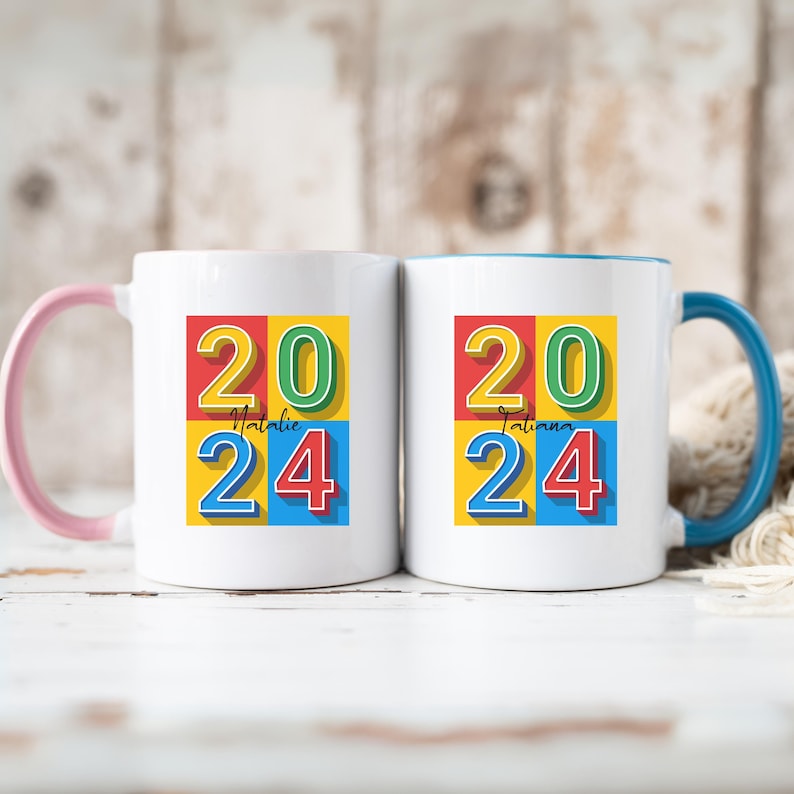 May include: Two white ceramic mugs with pink and blue handles. The mugs have a colorful design with the numbers 2024 in a retro style. The names "Natalie" and "Tatiana" are written on the mugs.