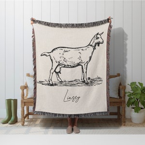 Pu&ograve; includere: Una coperta in tessuto intrecciato color crema con un'illustrazione in bianco e nero di una capra e il nome "Lindsey" scritto in corsivo. La coperta ha frange sui bordi.