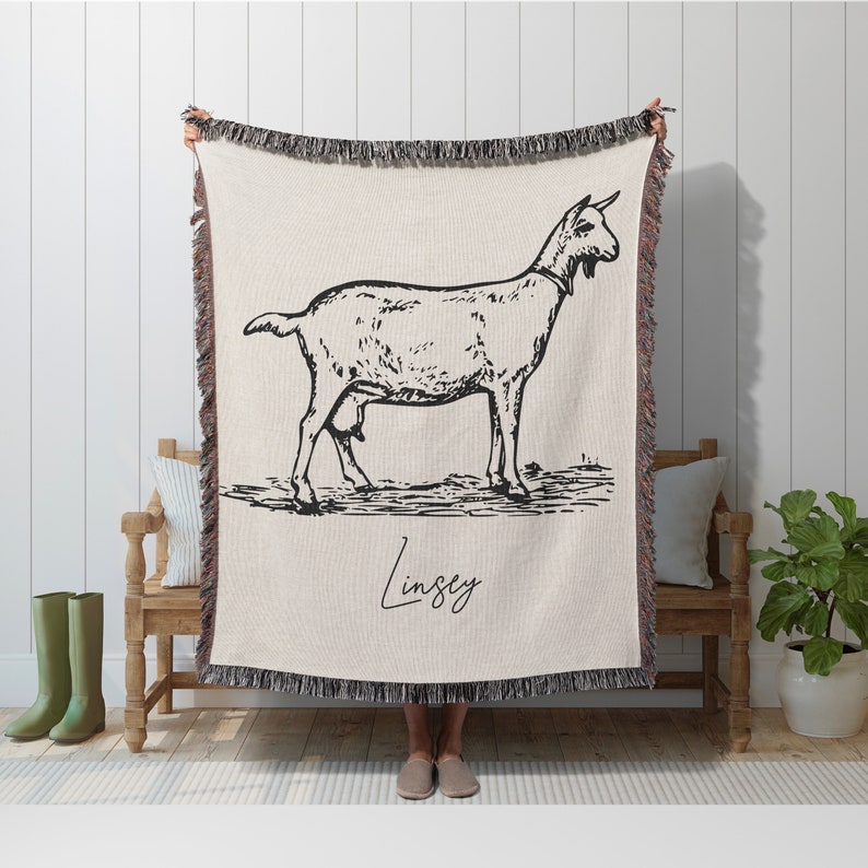 Pu&ograve; includere: Una coperta in tessuto intrecciato color crema con un'illustrazione in bianco e nero di una capra e il nome "Lindsey" scritto in corsivo. La coperta ha frange sui bordi.