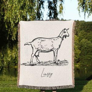 Pu&ograve; includere: Una coperta in tessuto intrecciato con un'illustrazione in bianco e nero di una capra. La coperta ha un bordo a frangia e il nome "Lindsey" &egrave; scritto in corsivo in basso.