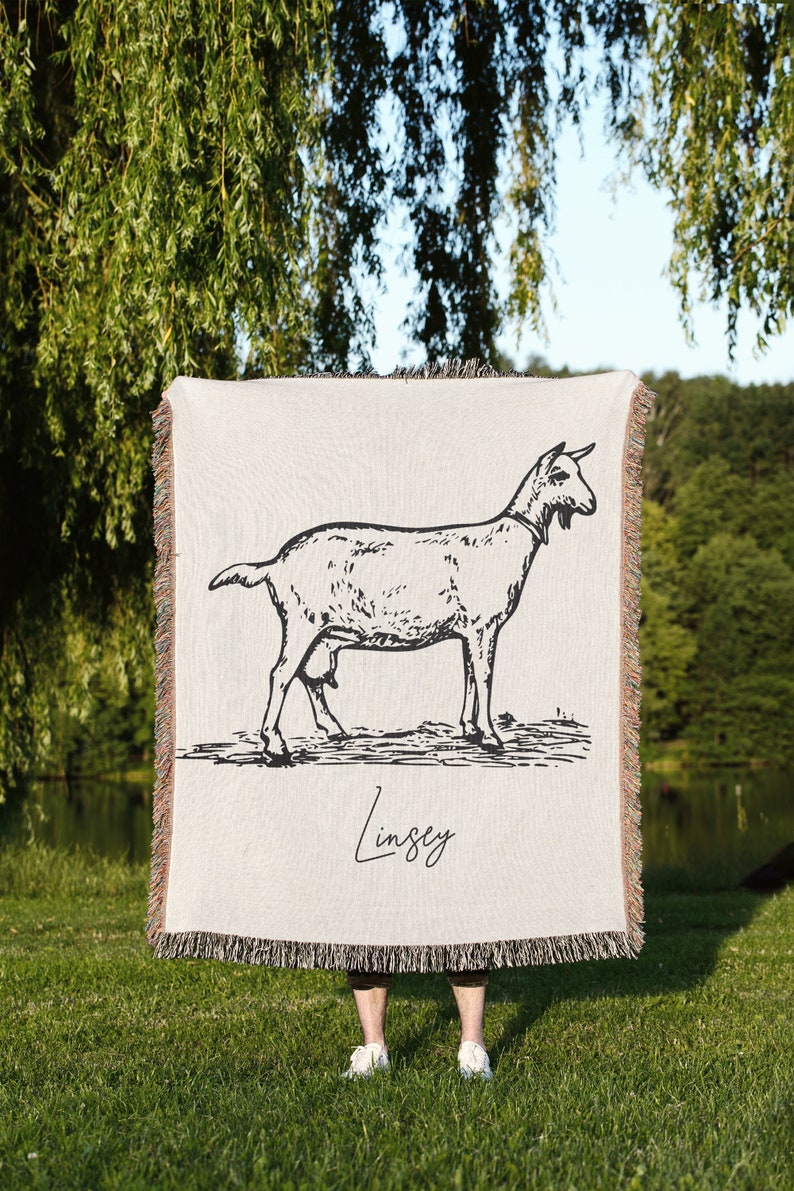 Pu&ograve; includere: Una coperta in tessuto intrecciato con un'illustrazione in bianco e nero di una capra. La coperta ha un bordo a frangia e il nome "Lindsey" &egrave; scritto in corsivo in basso.