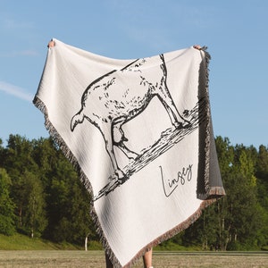 Pu&ograve; includere: Una coperta in tessuto bianco con un'illustrazione in bianco e nero di una capra e il nome "Lindsey" scritto in corsivo nero.