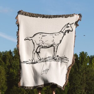 Pu&ograve; includere: Una coperta in tessuto bianco con un'illustrazione in bianco e nero di una capra. La coperta ha un bordo con frange e il testo "Lindsey" &egrave; stampato in nero nella parte inferiore.