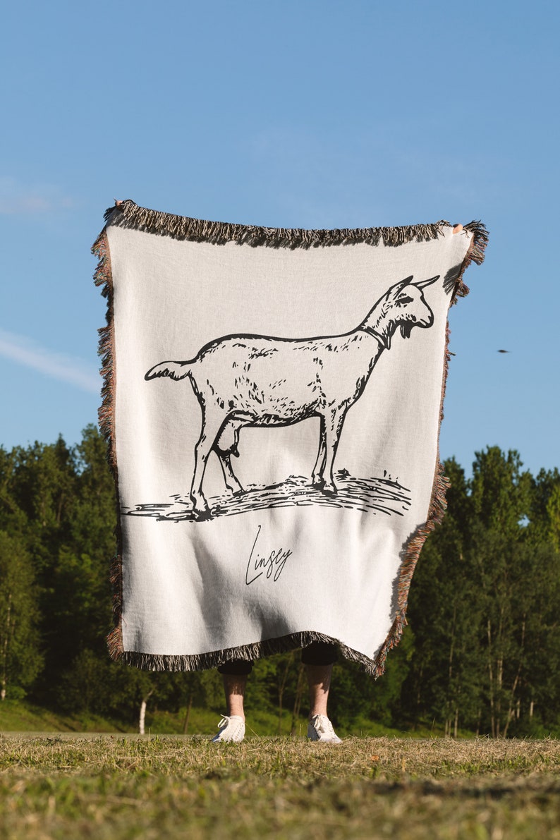 Pu&ograve; includere: Una coperta in tessuto bianco con un'illustrazione in bianco e nero di una capra. La coperta ha un bordo con frange e il testo "Lindsey" &egrave; stampato in nero nella parte inferiore.