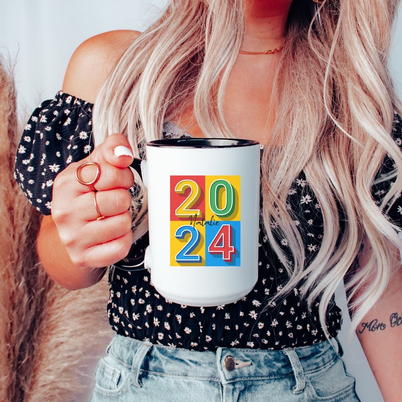 May include: White ceramic mug with a black rim and handle. The mug has a colorful design with the numbers 2024 in a block style font. The numbers are in red, yellow, blue, and green squares. The word "Natalie" is written in black cursive script above the number 24.