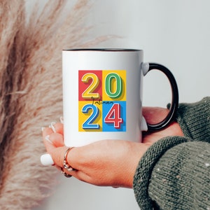 May include: White ceramic mug with a black handle. The mug has a colorful design with the numbers 20 and 24 in a block style font. The text "Tatiana" is written in cursive between the numbers.