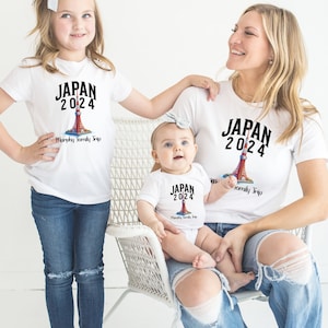 K&ouml;nnte beinhalten: Eine dreik&ouml;pfige Familie tr&auml;gt wei&szlig;e T-Shirts mit einem Motiv einer roten und wei&szlig;en Turms und dem Text "JAPAN 2024" sowie den Worten "Murphy Family Trip". Die Shirts sind auf wei&szlig;em Hintergrund.