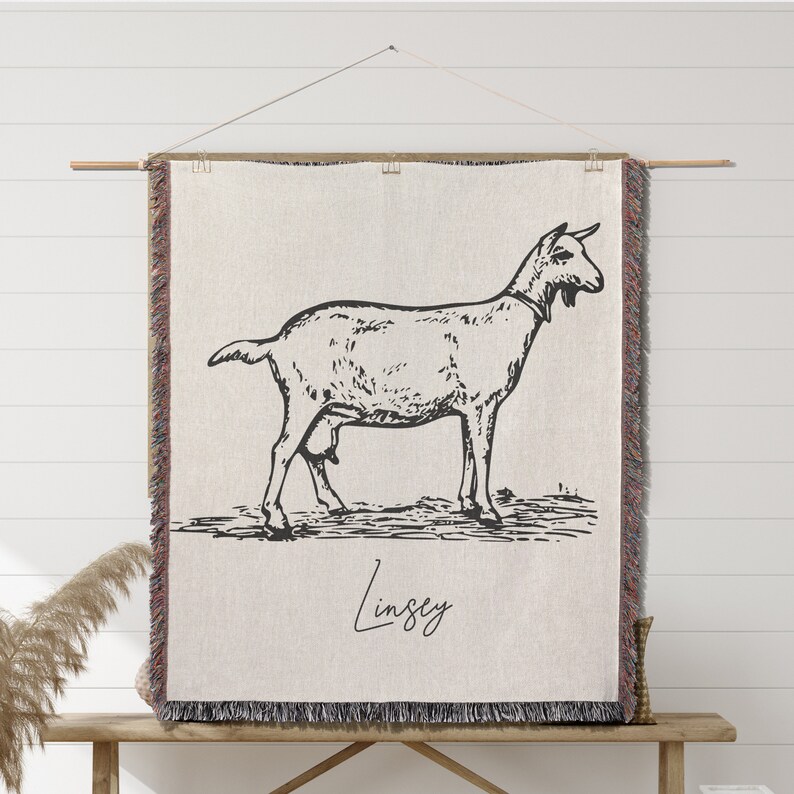 Pu&ograve; includere: Un arazzo da parete tessuto con un disegno in bianco e nero di una capra con il nome "Lindsey" scritto sotto. L'arazzo ha uno sfondo beige e un bordo con frange marroni.