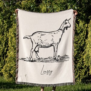 Pu&ograve; includere: Una coperta in tessuto di stile arazzo con un'illustrazione in bianco e nero di una capra e il nome "Lindsey" scritto in corsivo.