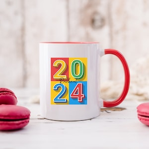 May include: White ceramic mug with a red rim and handle. The mug has a colorful design with the numbers 2024 in a block style font. The word "Rachel" is written in black script font below the numbers.