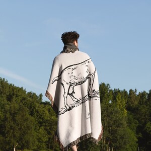 Pu&ograve; includere: Una persona indossa una coperta bianca con un'illustrazione in bianco e nero di una capra. La coperta ha frange sui bordi. La persona cammina in un campo erboso.