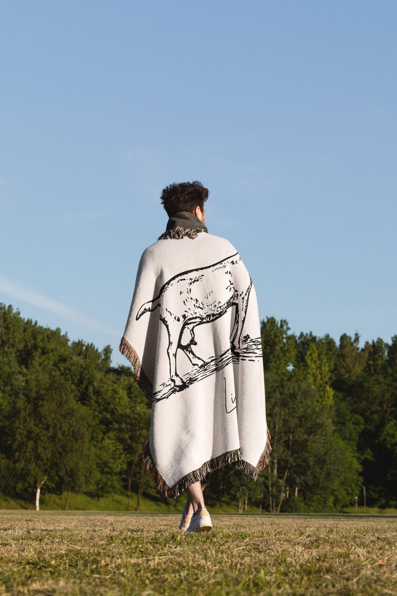 Pu&ograve; includere: Una persona indossa una coperta bianca con un'illustrazione in bianco e nero di una capra. La coperta ha frange sui bordi. La persona cammina in un campo erboso.