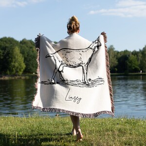 Pu&ograve; includere: Una coperta in tessuto bianco con un disegno a linea nera di una capra e il nome "Lindsey" stampato su di essa. La coperta &egrave; tenuta da una persona in piedi davanti a un lago.