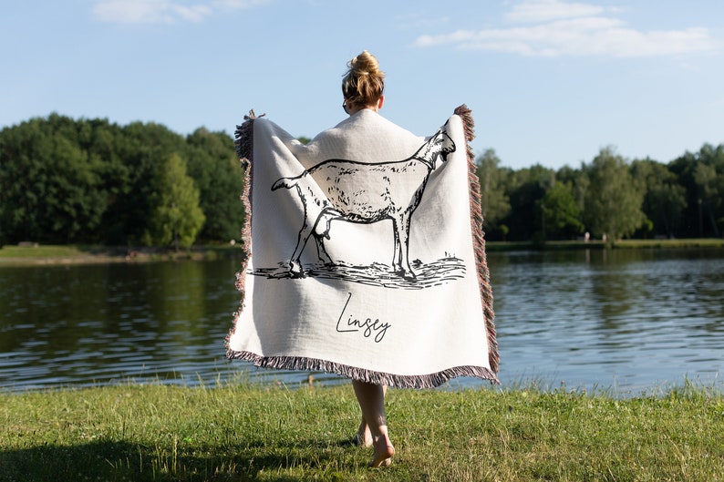 Pu&ograve; includere: Una coperta in tessuto bianco con un disegno a linea nera di una capra e il nome "Lindsey" stampato su di essa. La coperta &egrave; tenuta da una persona in piedi davanti a un lago.