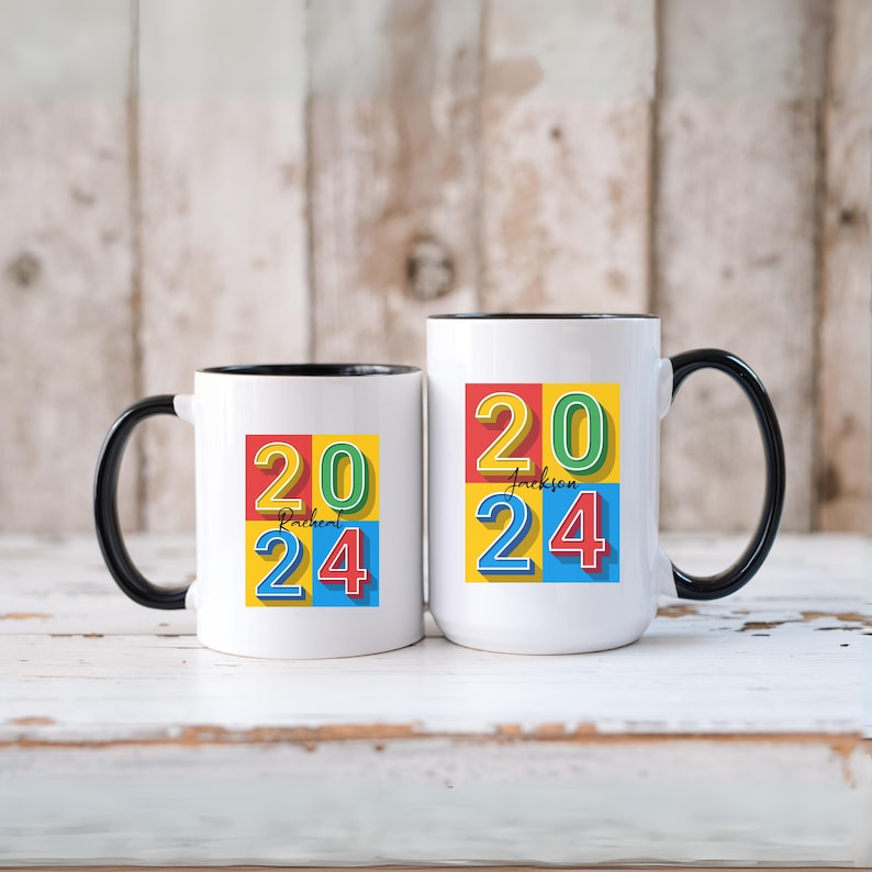 May include: Two white ceramic mugs with black handles and black rims. The mugs have a colorful design with the numbers 20 and 24 in a retro style. The mugs are on a white wooden surface.