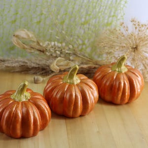 May include: Three orange ceramic pumpkins with green stems sit on a wooden surface. The pumpkins have a ribbed texture and are arranged in a row. Dried plants and a green mesh background are visible.