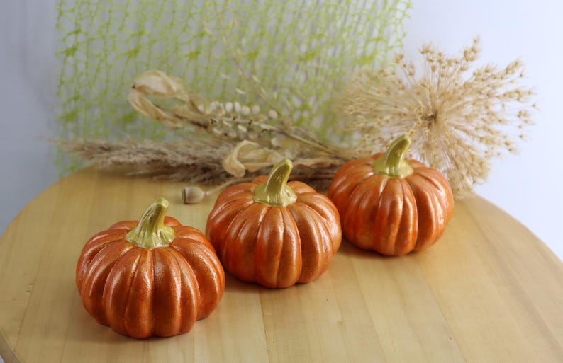 May include: Three orange ceramic pumpkins with green stems sit on a wooden surface. The pumpkins have a ribbed texture and are arranged in a row. Dried plants and a green mesh background are visible.