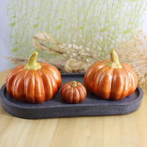 May include: Three orange ceramic pumpkins with green stems sit on a dark gray oval tray. The pumpkins vary in size, with the largest on the left and right, and a smaller one in the center. The pumpkins are on a wooden table.