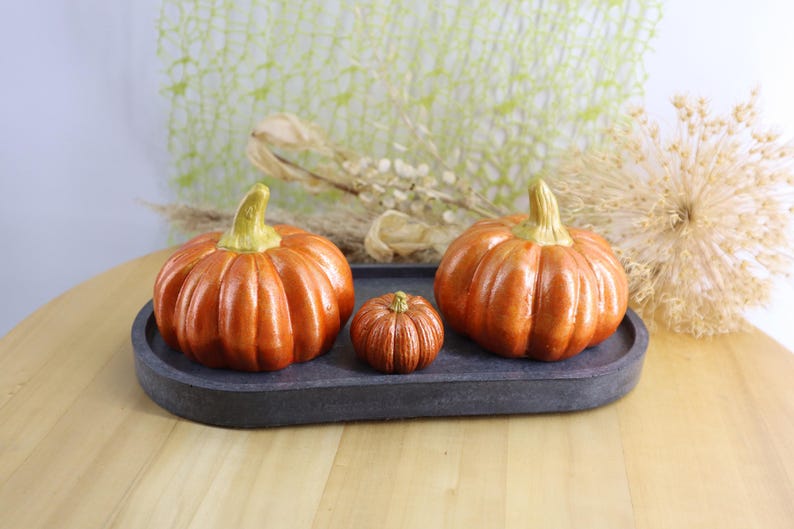 May include: Three orange ceramic pumpkins with green stems sit on a dark gray oval tray. The pumpkins vary in size, with the largest on the left and right, and a smaller one in the center. The pumpkins are on a wooden table.