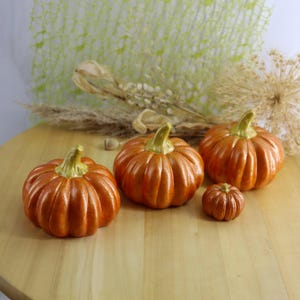 May include: Four orange ceramic pumpkins of varying sizes are arranged on a wooden surface. The pumpkins have green stems and are a glossy orange color. The smallest pumpkin is a darker shade of orange.
