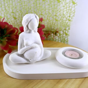 May include: A white sculpture of a pregnant person seated in a meditative pose, on a white tray. A white, oval-shaped candle holder with a pink candle is next to the sculpture. Red and green flowers are in the background.