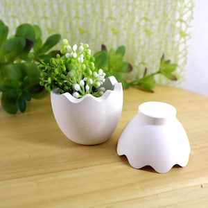 Könnte beinhalten: Ein weißer, eiförmiger Pflanztopf mit gewelltem Rand, der eine kleine Anordnung aus grünen und weißen Kunstblumen enthält. Der Pflanztopf befindet sich neben seinem passenden Deckel, ebenfalls weiß mit gewelltem Rand. Auf einer Holzoberfläche.