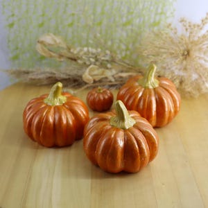 May include: Four orange ceramic pumpkins with green stems are arranged on a wooden surface. The pumpkins vary in size, with the largest ones in the background and the smallest in the center. Dried plants are in the background.