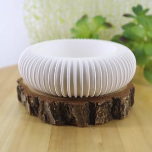 May include: A white, circular, ribbed planter with a hollow center and a rounded, donut-like shape. The planter features a series of vertical, evenly spaced ridges around its exterior, creating a textured, pleated appearance throughout the surface.