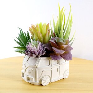 Könnte beinhalten: Ein weißer Keramik-Pflanztopf in Form eines Oldtimer-Vans, gefüllt mit bunten Kunst-Sukkulenten. Die Sukkulenten umfassen grüne, violette und gelb-grüne Sorten und bilden eine lebendige Darstellung. Der Pflanztopf hat detaillierte Merkmale wie Räder und Fenster.