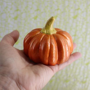 May include: A small, orange ceramic pumpkin with a green stem. The pumpkin is held in a hand, showcasing its ribbed texture and autumnal color. The background is a soft, patterned design, suggesting a seasonal theme.