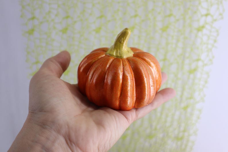 May include: A small, orange ceramic pumpkin with a green stem. The pumpkin is held in a hand, showcasing its ribbed texture and autumnal color. The background is a soft, patterned design, suggesting a seasonal theme.