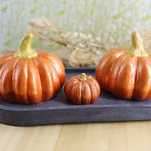 May include: Three orange pumpkins of varying sizes sit on a dark gray oval tray. The pumpkins have textured surfaces and green stems. The tray is on a light wooden surface, with a blurred background.
