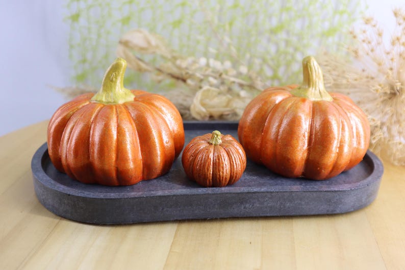 May include: Three orange pumpkins of varying sizes sit on a dark gray oval tray. The pumpkins have textured surfaces and green stems. The tray is on a light wooden surface, with a blurred background.