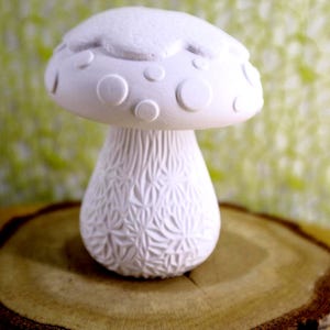 May include: A white ceramic mushroom figurine with a textured cap and stem. The cap features circular and crescent-shaped details. The stem has a detailed, geometric pattern. The figurine is displayed on a wooden slice.