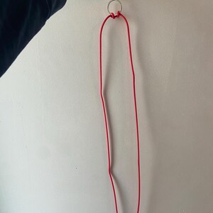 May include: A red lanyard with a silver metal ring and clasp. The lanyard is made of a thin, flexible material and is hanging against a white wall. The lanyard is held by a hand.